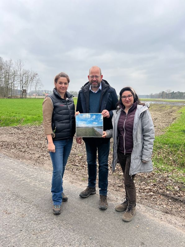 3 Personen stehen am Rande einer Naturfläche mit Grün- und Ackerbereichen und zeigen einen Geländeplan in die Kamera